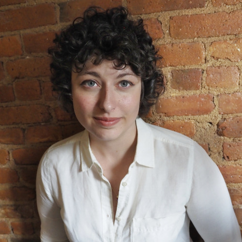 Robyn with curly brown hair and white shirt standing in front of brick wall